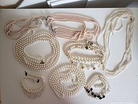 Top view of nine pearl necklaces and one bracelet laid out on a table showing variety in pearl size, strand number, and bead color.