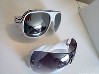 Chunky white framed sunglasses with mirrored lenses and wraparound gradient lens sunglasses with white frame laid on a flat surface.