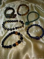 Top-down view of six assorted bracelets arranged on a gold satin fabric. Bracelets vary in material, including metal and various beads in purple, teal, black, and brown tones.