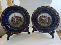 Two porcelain plates with cobalt blue and gold leaf rims on black stands, showing painted Napoleon battle scenes central on each plate.