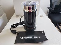 Full view of the Nespresso Aeroccino3 milk frother with power cord and included user manual on a white table.