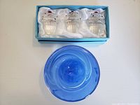 View of three cupcake-shaped glass wine stoppers in blue padded box with white satin lining and blue Kosta Boda votive candle holder on plain surface.