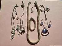Four sets of vintage costume jewelry including necklaces, bracelets, and earrings displayed on a flat surface with white background. Jewelry pieces feature silver and gold tone metals with various shaped faux stones in green, purple, and blue hues.