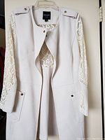 Front view of the off-white long jacket showing crochet lace sleeves, epaulets, and front pockets with buttons.
