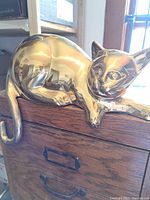 Side view of polished brass cat sculpture perched on wooden shelf edge, showing reflective surface and curled tail.