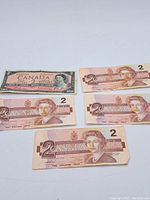 Photo showing five 1986 series Canadian $2 bills with Queen Elizabeth II portrait and one vintage $2 bill with pink tone and older design.