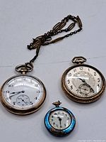 Three pocket watches displayed on a white surface: two larger gold watches and one smaller blue watch