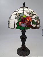 Full view of Tiffany style stained glass lamp illuminated showcasing rose floral pattern on shade and heavy metal base with carved details.