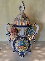 Front view of large ceramic lamp with floral decoration and three-dimensional flowers attached to sides, showing light glowing through cutouts.