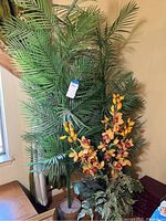 Photo showing tall faux palms, ferns, and yellow/magenta artificial flowers in a large vase, arranged against a beige wall.