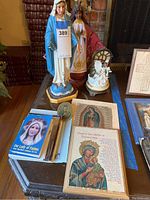 Photo shows two religious statues, a glass dome figurine with angels, and several prayer cards and framed art on a table.