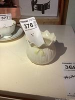 Front angle view of cream-colored decorative bowl with ruffled rim and ridged body on table with auction tag.