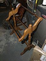 Front angle view of the solid wood blanket rack showing the curved side panels, three horizontal dowels, and rounded feet on a concrete floor.