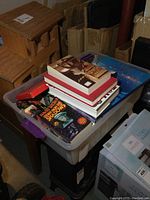 Plastic bin filled with multiple books stacked inside, including some large and small volumes, with a neck magnifier visible among the items.