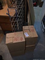 Photo showing four cardboard cases stacked with empty green glass wine bottles inside compartments and the metal wine rack behind them.