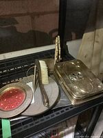 Photo showing brass smoker's box, crumb brush with handle, and round sundial tray arranged on shelving.