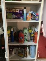 White shelving unit with various cleaning supplies like sprays, liquids, and powders including Vim, Windex, and other brands, along with a box of plastic bags and small plastic storage unit.