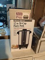 Two stacked boxes, larger box is West Bend 12 to 30 Cup Party Perk Coffee Urn, smaller box on top is Spring Gourmet Line warming grill box showing brand and product names.