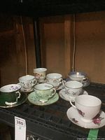 Photo showing eight teacups and saucers with varying floral patterns and colors on a black shelf.