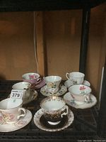 Seven sets of vintage decorative cups and saucers on shelving, various floral designs and colors visible