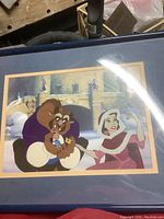 Framed lithograph of Beast and Belle in winter attire, holding blue bird in castle courtyard setting. Reflections and wear visible on plastic protective cover.