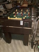 Foosball table front and side view showing dark wood cabinet, legs, and ball return slot.