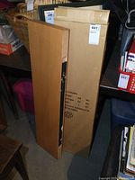 Two IKEA wall shelves, one boxed, one unboxed, standing vertically against a shelf with surrounding items