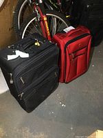 Photo of two wheeled carry-on suitcases, black and red, standing upright on floor near bicycle and U-Haul box.