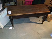 Full view of antique wood piano bench showing rectangular top and four legs on concrete floor with visible wear.