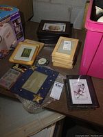 Overall collection of picture frames including stacks of dark wood IKEA frames, stacks of light wood 5x7 frames, a decorative blue star frame, and new 4x6 frames along with small items on table.