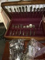 Solid wood canteen open showing partial silverplate flatware set inside and sealed plastic bags of mixed stainless steel flatware on table in front