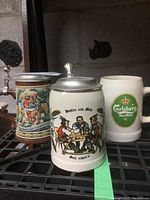 Photo of three beer drinking vessels on shelf: Carlsberg mug and two lidded German steins.
