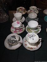 Photo showing all 8 matching teacups and saucers with various floral and scenic designs on a dark surface.