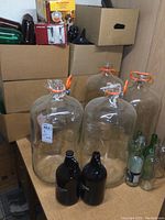 Four large clear glass carboys with orange sealing clamps on table with cardboard boxes containing other wine making supplies