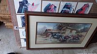 Photo showing framed art print with vintage cars parked near an old building in countryside, framed in brown wood.