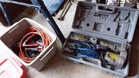 Plastic case open showing electric car jack with wired remote control, battery charger cables with alligator clips coiled inside a plastic tub to the left.