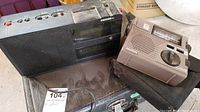 Proton 320 AM/FM alarm clock radio in black with buttons and digital display shown next to a brown Grundig 200 emergency radio with tuning dial and speaker grill, both placed on a surface.