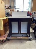 Full front view of the bathroom vanity with two dark wood doors with frosted glass panels and white rectangular sink on top. The vanity is placed on a trolley.