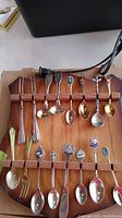 Overall front view of wooden display board holding 17 assorted collector spoons with various designs and colors on handles.
