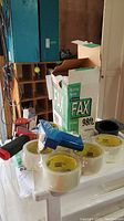 Photo of 6 rolls of clear packing tape, blue labeler tool, and fax paper box in garage setting.