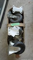 Photo showing several heavy-duty dolly wheels stacked inside a wooden box, featuring metal mounting plates and large black rubber wheels.