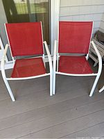 Photo showing two identical outdoor chairs with white metal frames and red cloth seating and backrest, placed on a wooden deck against a home exterior wall.