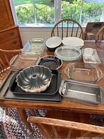 Full view of assortment of bakeware including Bundt pan, cookie sheets, loaf pans, glass dishes, and pie plates on wooden table near window.