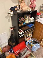 Black bookshelf containing various items including dolls, books, boxes, and sports equipment in and around it.