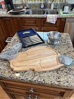 Photo of entire lot on granite countertop under kitchen window showing all items: six cheese knives in box, rabbit-shaped wooden board with metallic rabbit head and tail, two silver-tone metal dipping bowls.