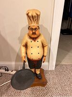 Chef statue figurine with beige chef hat, glasses, mustache, holding frying pan, standing on wooden base, carpet and wall background
