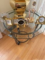 Seven assorted decorative table clocks arranged on a glass-top table with a brass base, showing different styles and sizes, mostly brass with some glass and black elements.