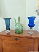 Three colored glass pieces displayed on a wooden cabinet against a white wall: two blue vases and one green bottle with cork stopper.