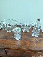 Four cut glass items on wooden surface: scalloped edge bowl, pedestal bowl, covered jar, and whiskey decanter with stopper.