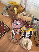 Overview of all knitting and yarn supplies arranged on floor including bags, box of yarn, and containers.